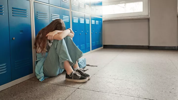 Teenager with light skin tone sits on the floor against a row of lockers with her head in her arms.