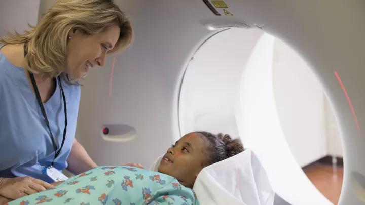 Child with a medium-dark skin tone wearing a hospital gown lays on a magnetic resonance imaging (MRI) machine indoors and smiles up at a technician with a light skin tone wearing blue scrubs and an ID badge.