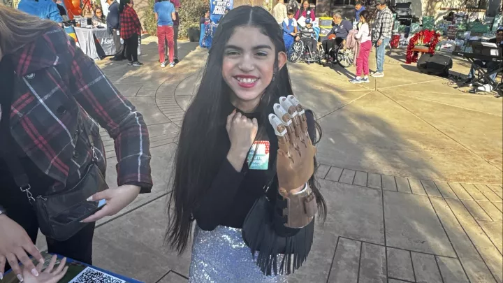 A child with medium light skin tone smiles and wears a 3D printed prosthetic hand.
