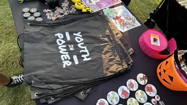 Overhead photo of a table with drawstring bags, buttons, stickers and other giveaway materials labeled with a “Youth in Power” logo at an outdoor event with two people in casual clothing standing near it.