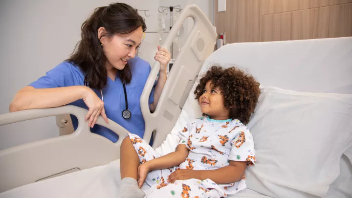 A dark skin-toned child in a hospital bed and a medium-light skin-toned woman in scrubs with a stethoscope around her neck smile at each other.