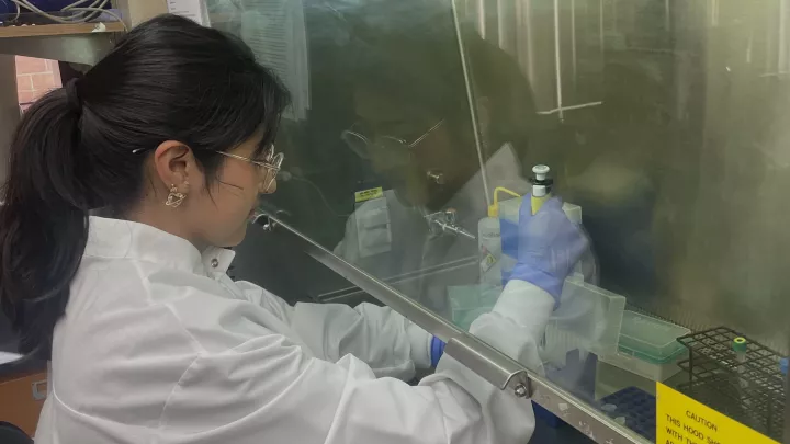 A young adult with a medium skin tone wears a white lab coat, glasses, and gloves and sits in a laboratory, holding various instruments used in medical research.