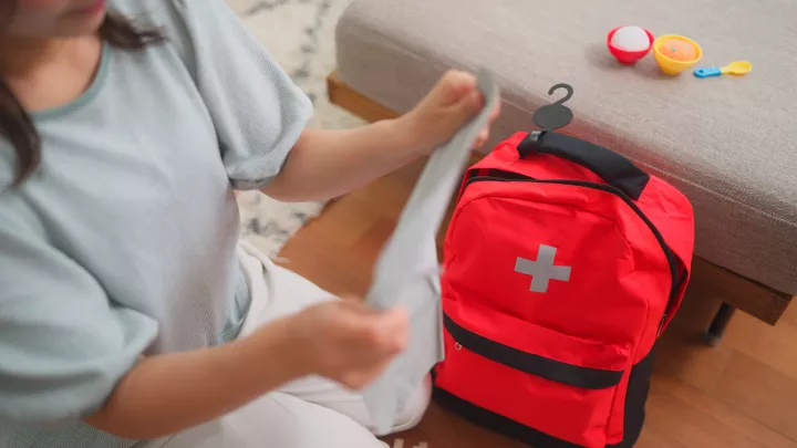 An adult with a medium light skin tone kneels next to a couch with baby food and dishes on it, folding a piece of clothing next to a fluorescent orange backpack with a silver emergency cross symbol on it.