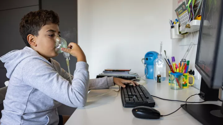 A child with medium skin tone holding an oxygen mask up to his face while sitting at a desk indoors and using a computer.