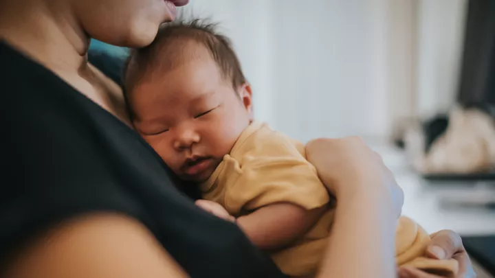 A medium skin-toned baby sleeps on the chest of a woman who is patting their back.