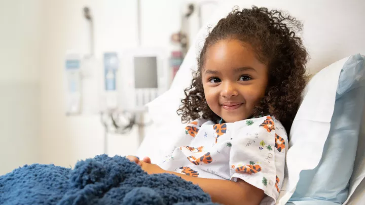 Young girl with medium skin tone and dark curly hair smiles from a hospital bed. She is wearing tiger pajamas and has a blue blanket over her lap.