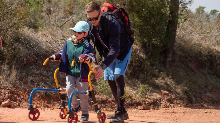 A child with a light skin tone uses a walker to walk along a dirt trail, while supported by an adult with a light skin tone.
