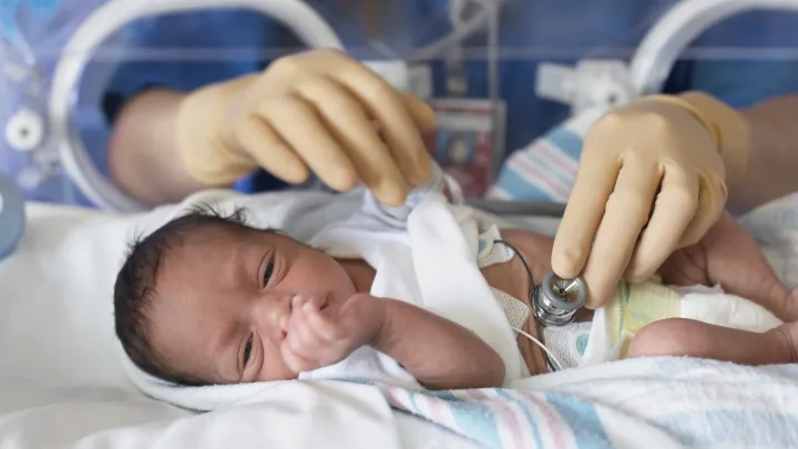 The gloved hands of a nurse in blue scrubs reach into an incubator, using a stethoscope to listen to a small newborn baby's heart.