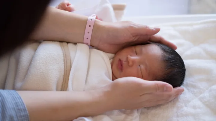 A newborn baby sleeps peacefully under a blanket, while a mother's hands gently cup the sides of the baby's head. One of the hands wears a pink plastic hospital bracelet. The mother's face is not shown.