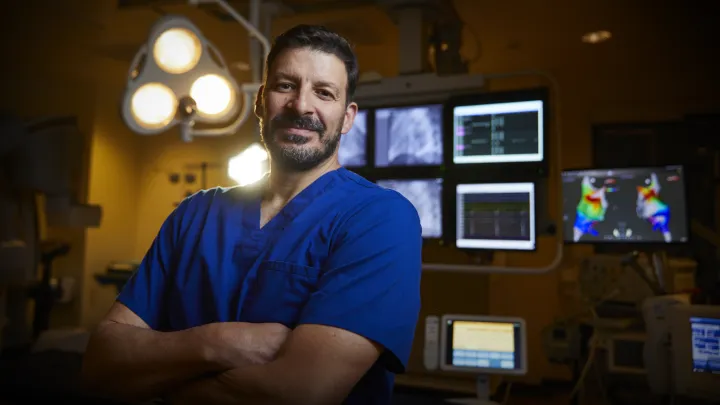 A man with medium skin tone and short dark hair and trimmed beard and mustache gazes at the camera with a slight smile. He wears blue scrubs, his arms are crossed, and medical lights and monitors are behind him.