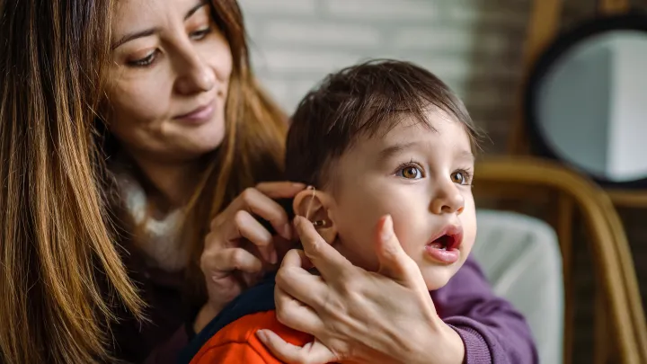 A mother sits behind her toddler son and adjusts his hearing aid. 