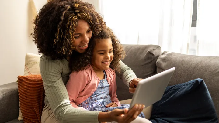 A little girl sits in her mom's lap on a gray sofa, smiling, as her mom points to something on a computer tablet. The mom and daughter have medium skin tone and shoulder length curly hair.