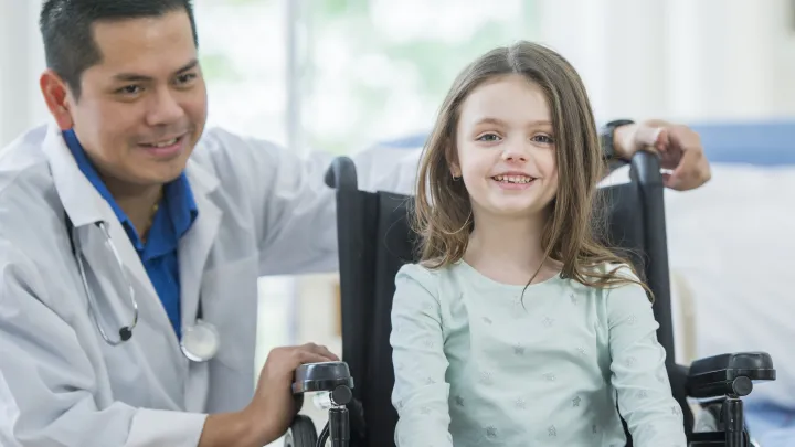 A young girl with light skin tone in a wheelchair smiles at the camera. A doctor with dark hair and medium skin tone and white coat is crouched behind her at her level, with his hands on the wheelchair.