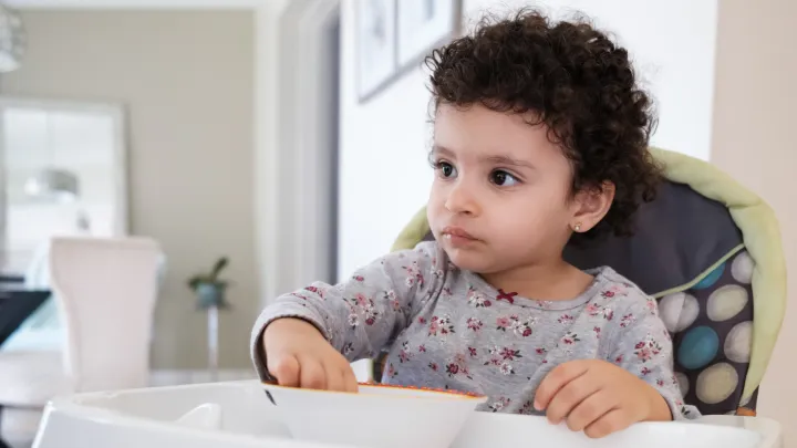A toddler sits in a high chair while hand-feeding themselves from a bowl.