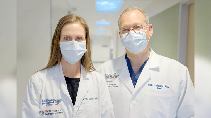A male and female doctor stand pose by side wearing white lab coats and blue medical procedure masks.