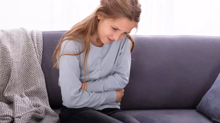 Young girl sitting on couch, holding stomach due to stomach pain