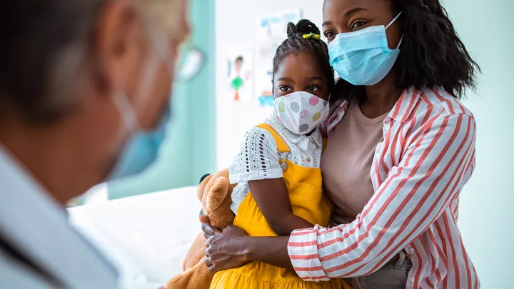 Woman wearing a blue medical procedure, holding a young girl, who is also wearing a medical procedure mask, while talking to a doctor.