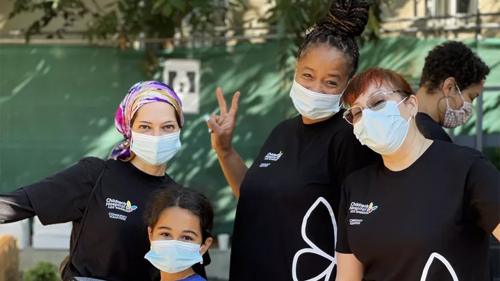 3 women in black CHLA T-shirts and a child in a blue shirt, all wearing surgical masks, smile at the camera. The medium-light skin-toned woman wears a colorful head scarf and the medium-dark skin-toned woman makes a piece sign with her fingers.  