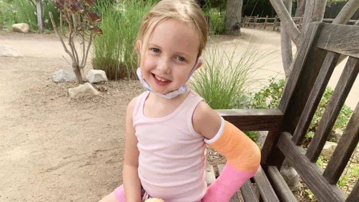 Little girl with light skin tone and left arm in a cast smiles as she sits on a wooden park bench