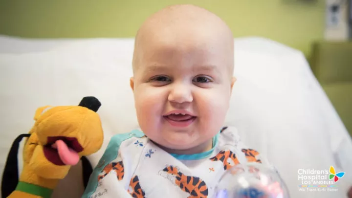 Young boy with light skin tone, shaved head and stuffed animal smiles as he sits in his hospital bed