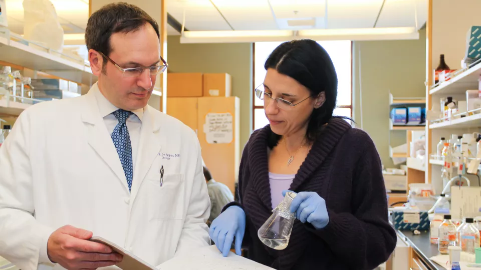 A woman with light skin tone holds a glass beaker covered with foil. She points into a notebook held open by a light skin toned man in a lab coat.