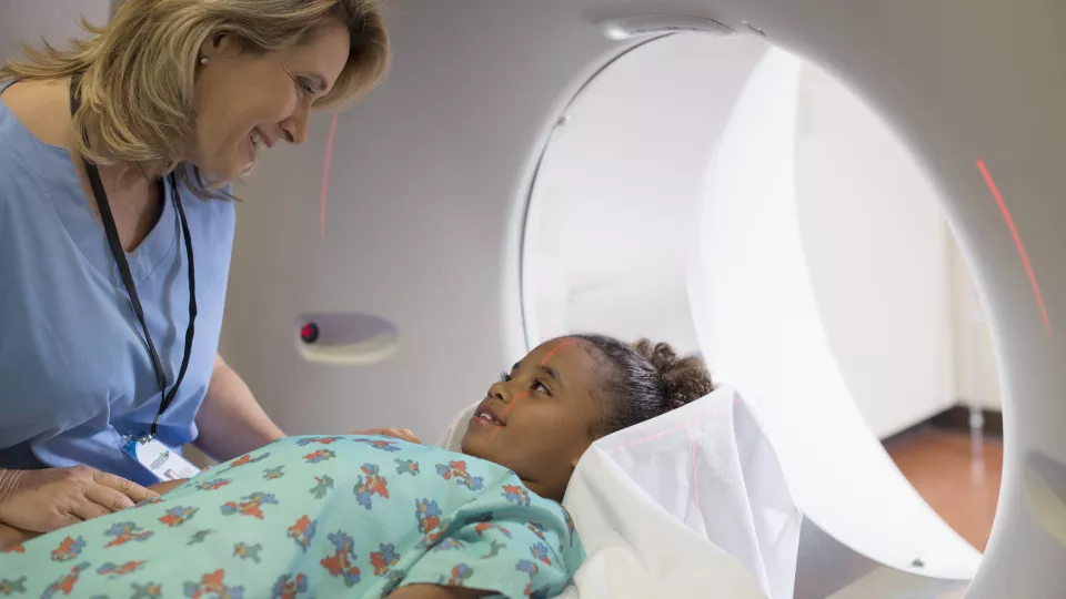 A woman with light toned skin wearing blue scrubs comforts a medium-dark skin toned girl laying on the bed of an MRI machine.