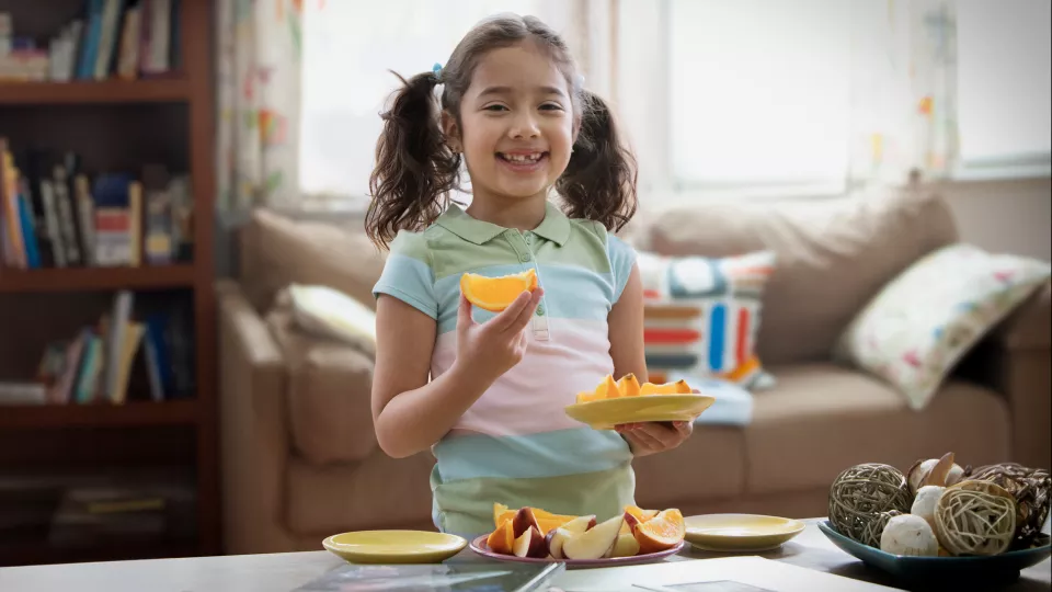 A girl with medium-light skin tone holds a plate of fruit and smiles. She has pigtails and a missing front tooth.