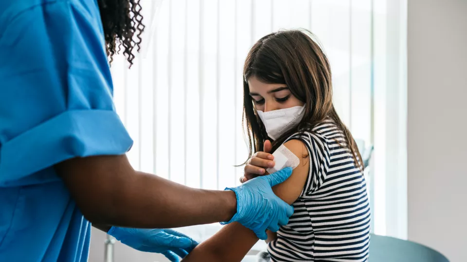 Female doctor with dark skin tone places bandage on upper arm of preteen girl with medium skin tone
