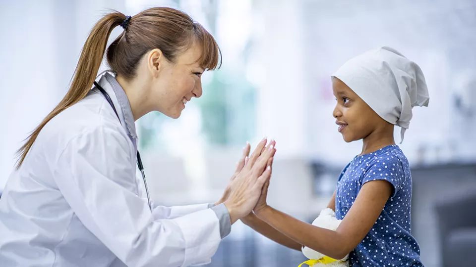 A female doctor with light skin tone smiles as she touches her palms to that of a young patient with dark skin tone and wearing a head scarf