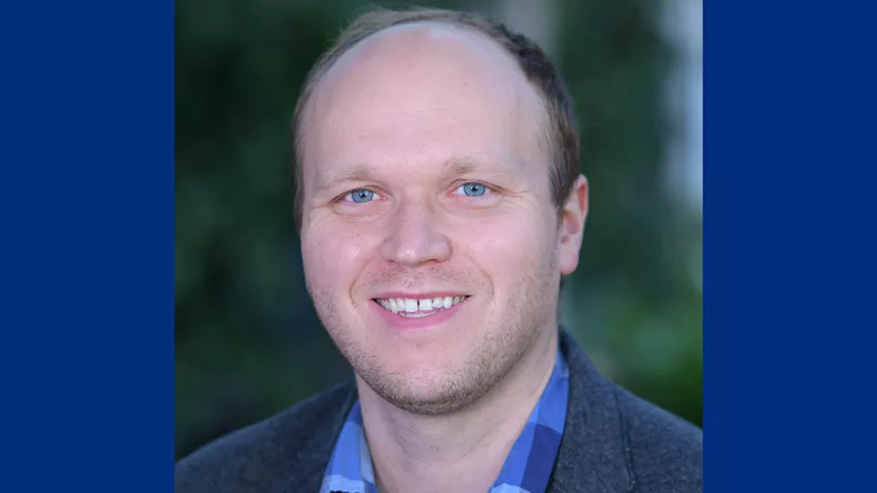 Headshot of a man with light skin tone and dark hair wearing a open collar shirt and blue blazer against a blurred outdoor background