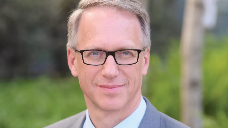Headshot of Dr. David Freyer, a man with light skin tone and light hair wearing glasses and business attire with green bushes in the background