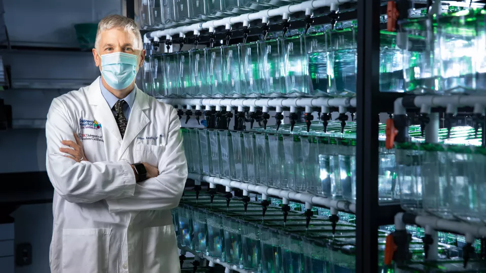 A researcher with light skin tone and grey hair wearing a surgical mask and white lab coat stands arms folded next to floor to ceiling shelving unit that holds row upon row of fishtanks