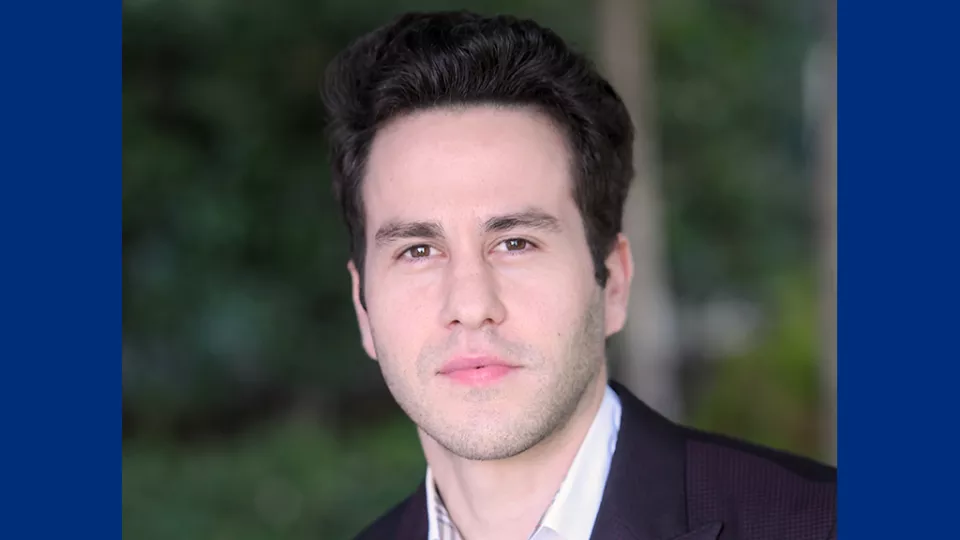 Headshot of a young man with light skin tone and dark hair wearing a white open-collar shirt and dark suit jacket against blurred outdoor background