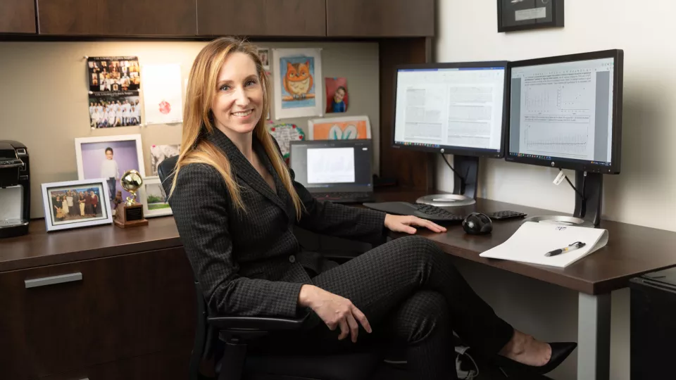 Smiling woman with light skin tone wearing dark business suit sits at desk with dual computer monitors in her office