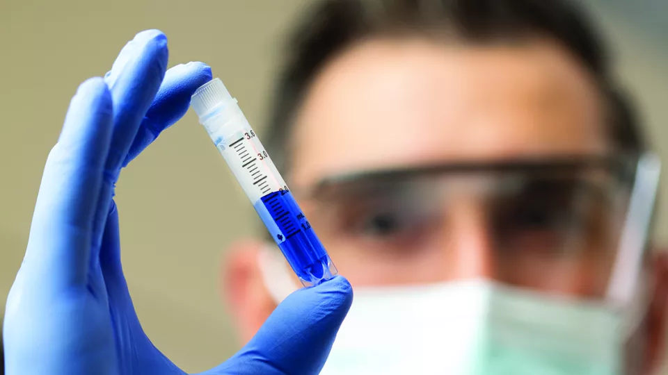 A male researcher with light skin tone wearing safety goggles, face mask and blue latex gloves holds a test tube containing a blue liquid