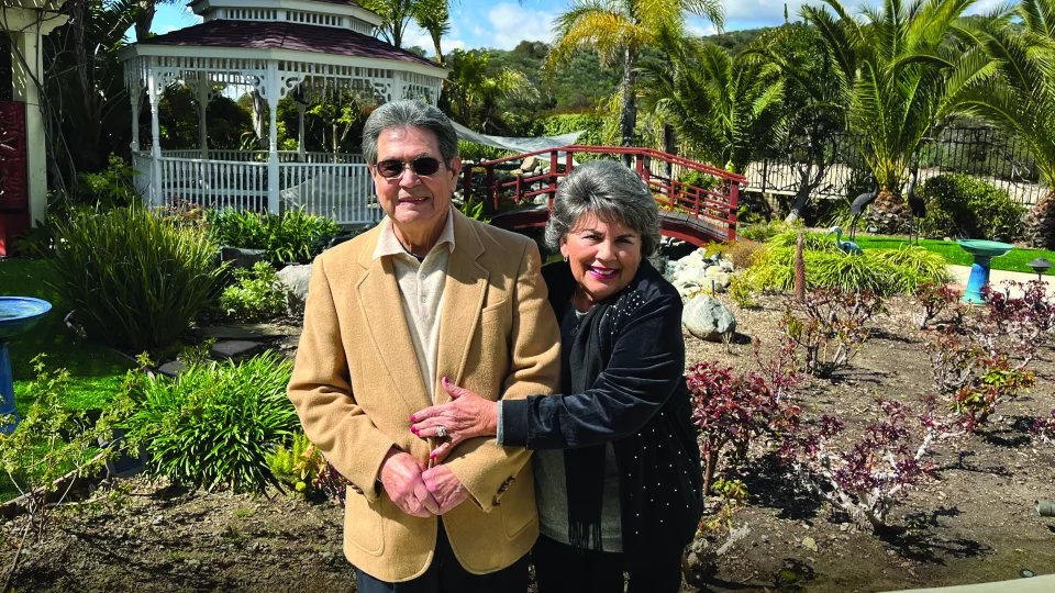 Older man and woman with light skin tone smile as they pose for a picture in front of a lush garden and gazebo