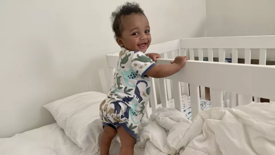 Standing infant with dark skin tone and dark hair wearing onesie with colorful dinosaur print smiles as they stand up supporting themselves on a crib railing
