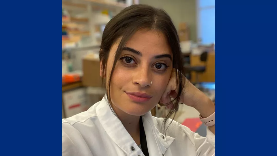 Headshot of a young woman with medium skin tone and dark hair wearing a white lab coat against a blurred laboratory background