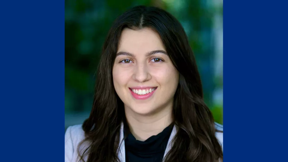 Headshot of a smiling woman with light skin tone and long dark hair wearing a dark blouse and light blue suit jacket against a blurred outdoor background