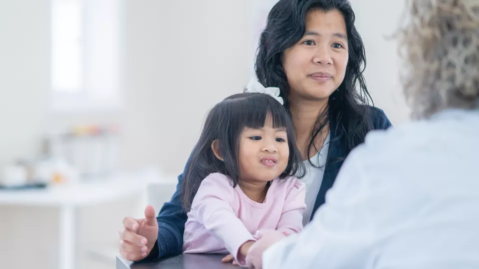 Young girl with medium skin tone and dark hair wearing purple top sits on lap of woman with medium skin tone and dark hair across from health care worker in doctor's office