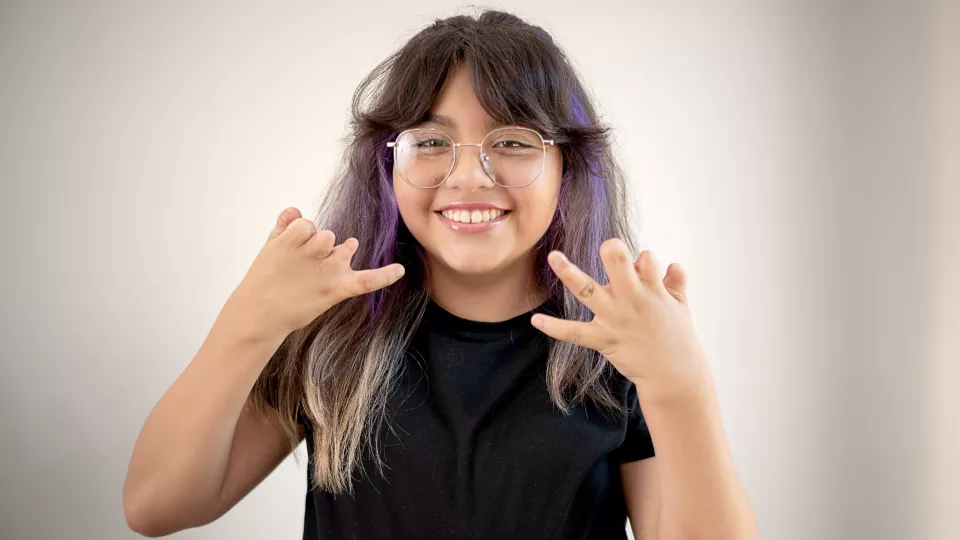 Smiling teenage girl with medium skin tone and dark hair with purple and blonde highlights wearing glasses and a black top shows off her hand differences against a neutral background