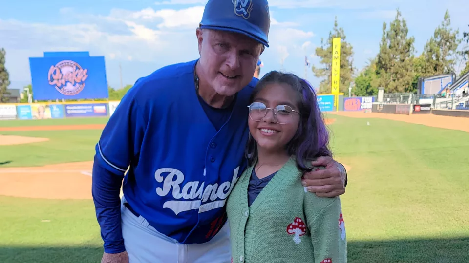 Middle aged man with light skin tone wearing baseball uniform poses with young girl with medium skin tone and glasses on baseball diamond