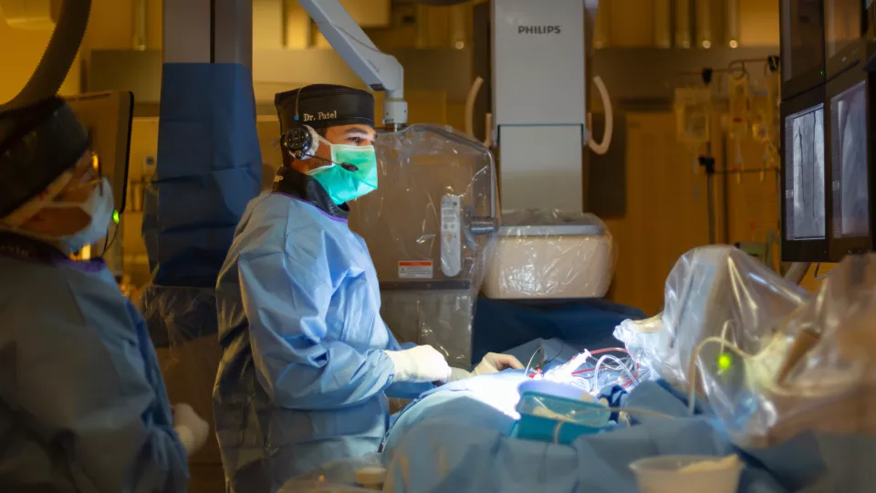 A masked male surgeon in blue surgical gown, green mask and black surgical cap, performs a cardiac catheterization procedure