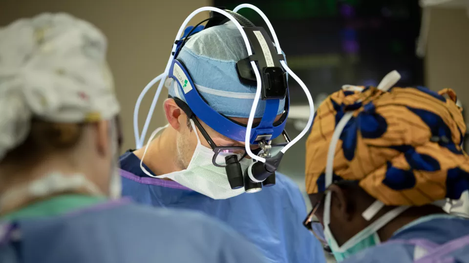 A close-up of Luke Wiggins, MD, during surgery with team members. He is wearing scrubs and a surgical headlamp and loupes