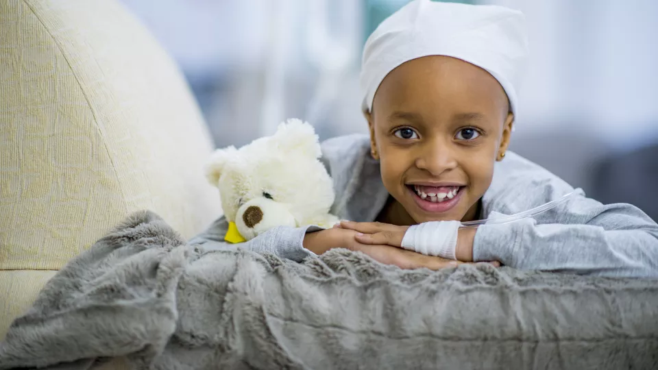 A little girl with dark skin tone and wearing a headscarf smiles as she lays in a hospital bed with her teddy bear cuddled close to her