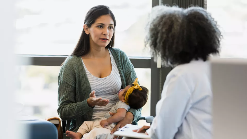 A mother with medium skin tone and dark hair holding a newborn baby girl gestures as she discusses her symptoms with a female doctor with dark hair and wearing a white lab coat