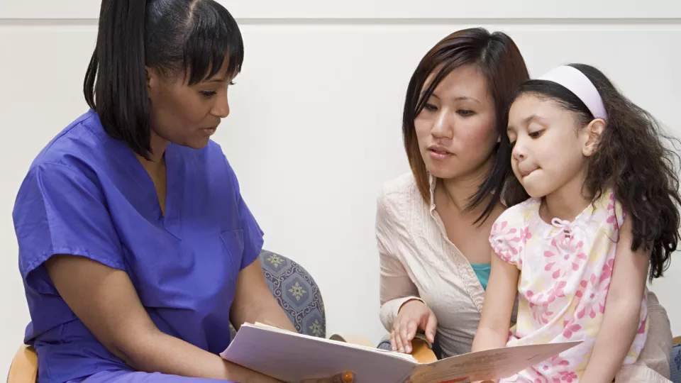 Nurse with dark skin and dark hair reviews chart with mother and daughter, both of whom have medium skin tones and dark hair