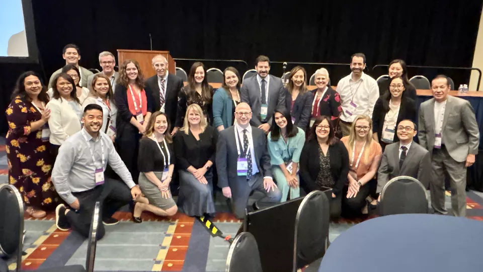 Two-dozen Heart Institute faculty in business attire pose in a hotel conference room, in front of a podium and stage.