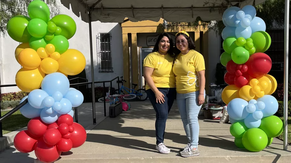 Two women with medium skin tone stand under a tent, between two pillars of colorful balloons.