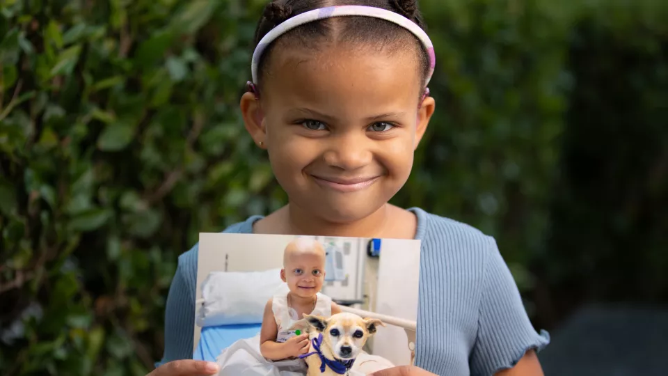 Young girl with medium skin tone and short dark hair wearing a blue top smiles as she holds up a photograph of herself as a younger cancer patient in a hospital bed with a small, light brown dog